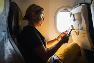 Passenger woman is flying in plane. Girl using phone sitting by airplane window. Traveling female with smartphone inside plane enjoying flight. Woman using wifi connection on board.