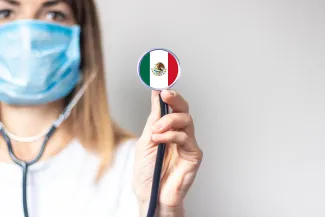 female doctor in a medical mask holds a stethoscope on a light background. Added flag of Mexico. 